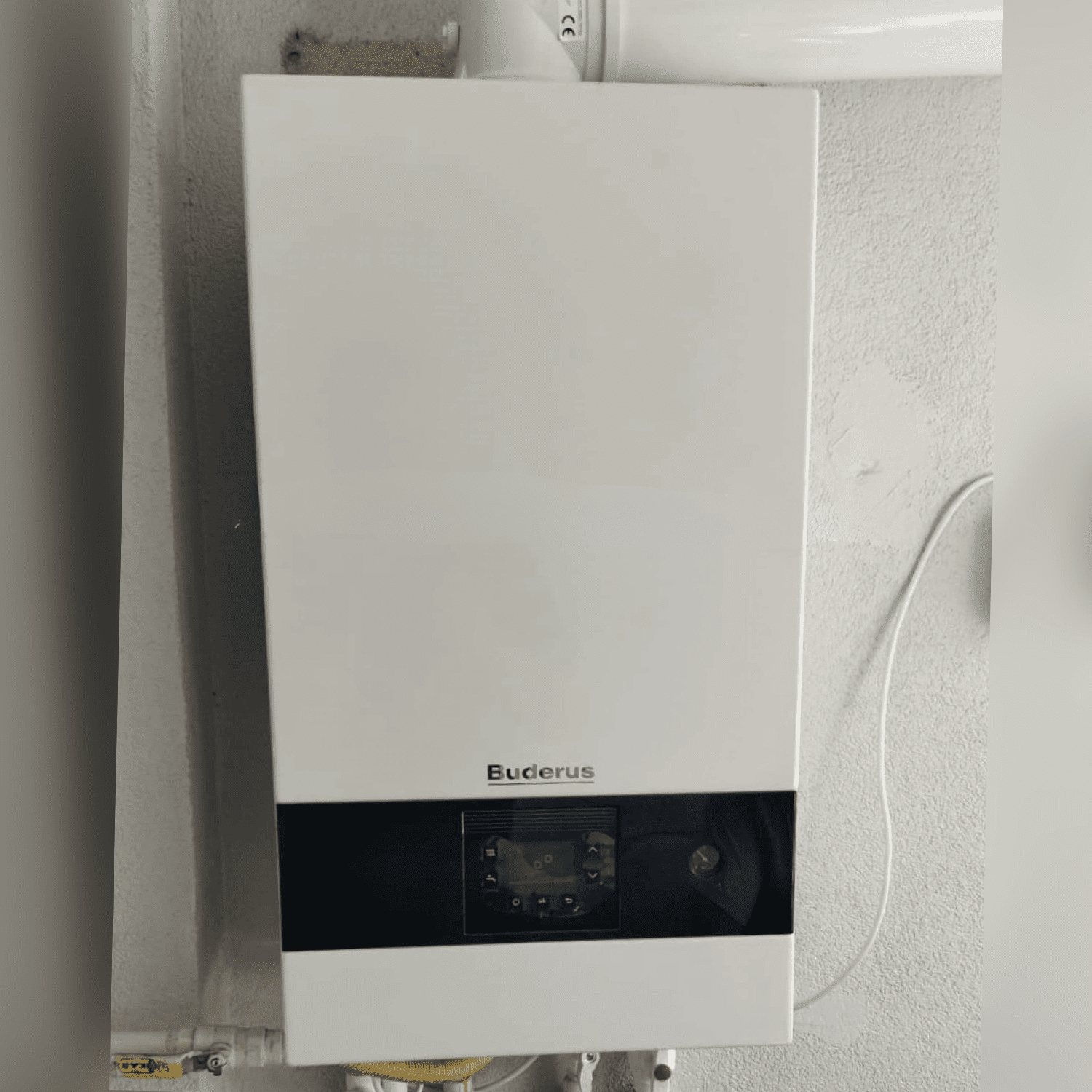 Beyaz duvara montajı yapılmış, alt tesisat boruları ve üst baca bağlantısı görünen Buderus Logamax plus GB122i.2 yoğuşmalı kombi fotoğrafı.